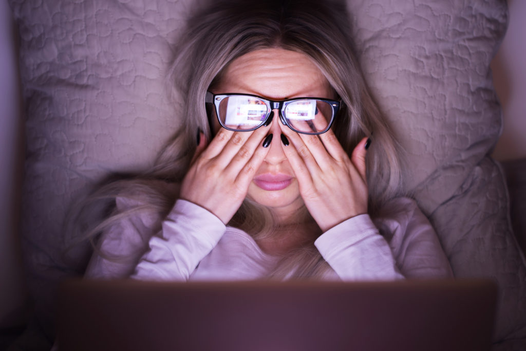 woman wearing glasses and rubbing her eyes while in bed looking at a laptop screen